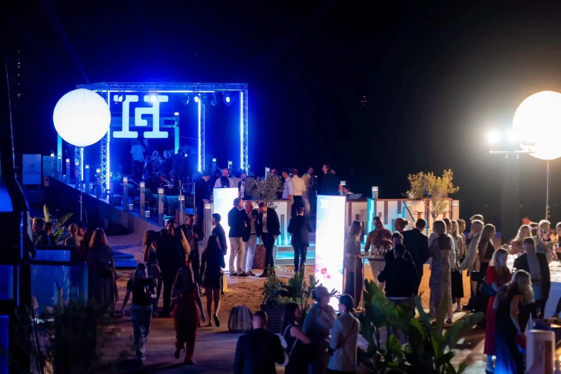 Guests walking along the beach in front of the lit Heinemann logo structure and glowing balloons, gala evening at night in Cannes.