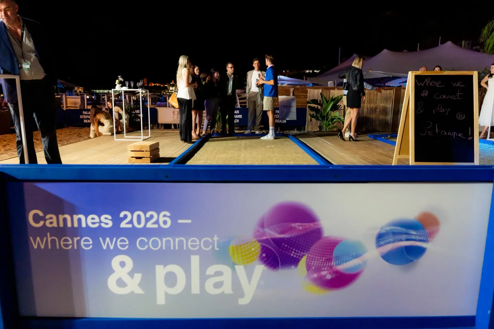 Cannes 2026 where we connect & play sign beside a boules court, guests chatting in the background, Heinemann gala evening.