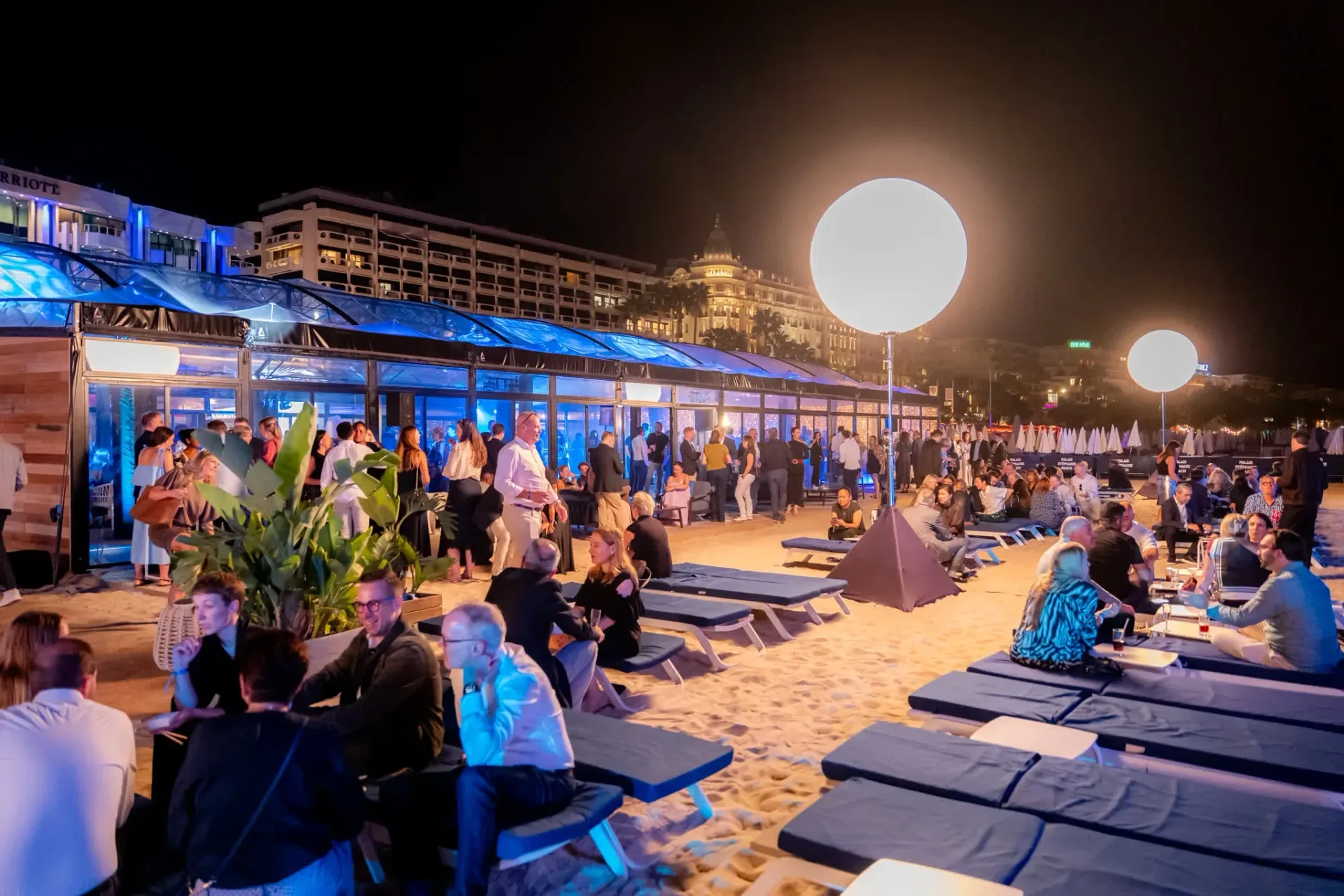 Guests relaxing on sun loungers and sand on the Cannes beach, Connect & Chill area, the illuminated Croisette behind, corporate gala evening.