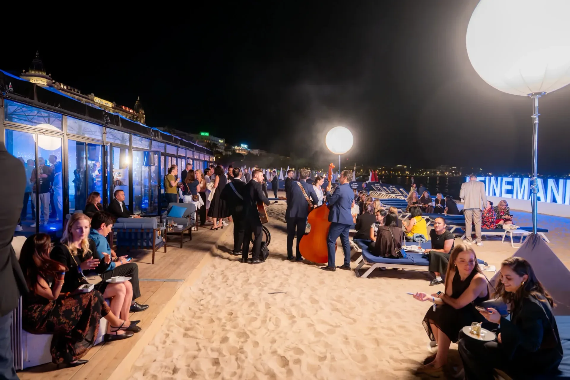 Invités installés sur le sable près des musiciens, zone Connect & Chill de la soirée de gala Heinemann à Cannes, lumière chaude et vue sur la baie.