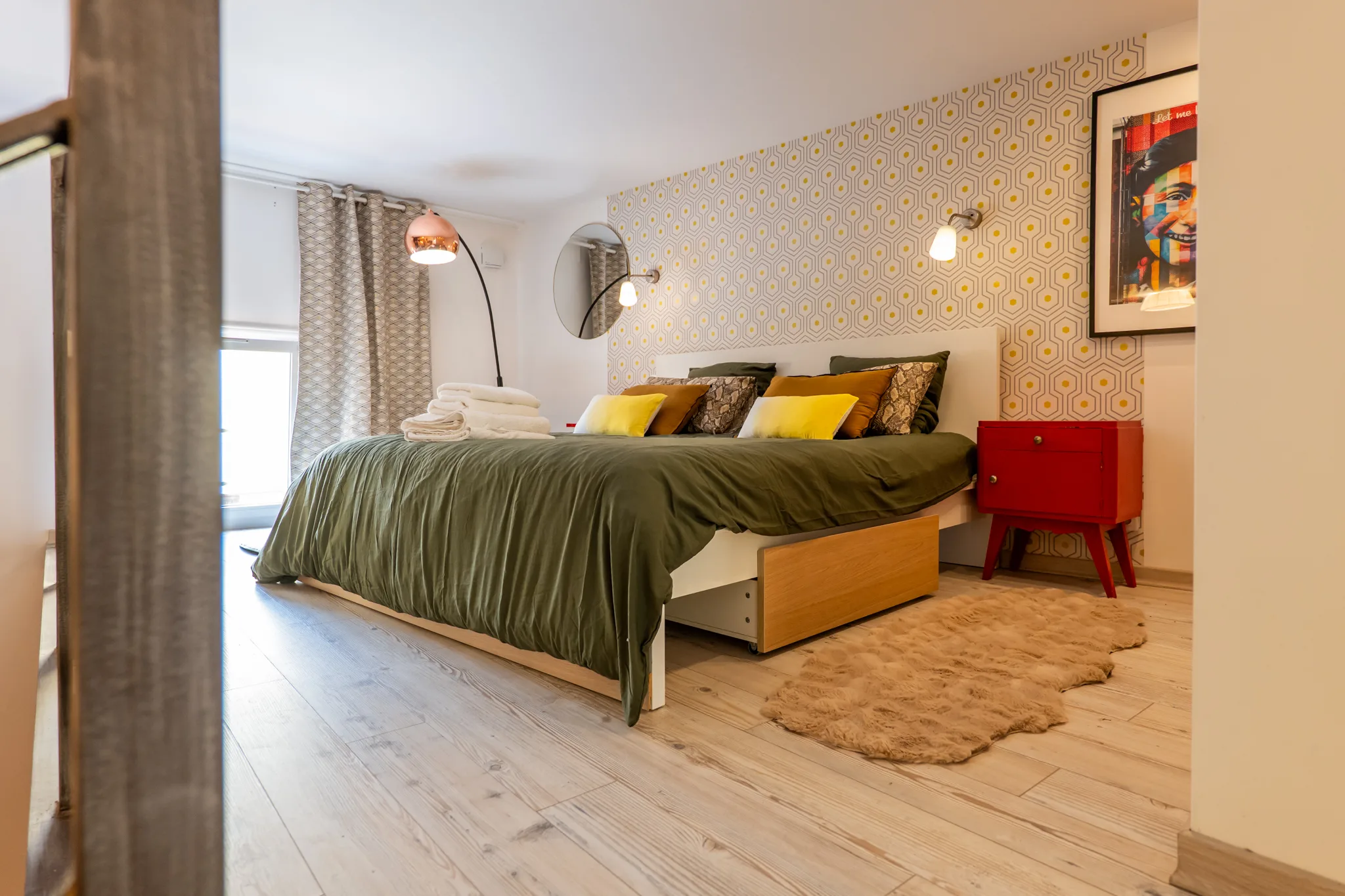 Lit avec coussins colorés (moutarde, jaune, python) et chevet rouge vintage dans la chambre en mezzanine.